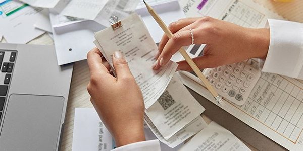 Unrecognizable woman holds receipts pencil uses laptop plans budget manages expenses calculates financial reports checks internet banking service poses at desktop with papers around. View from above